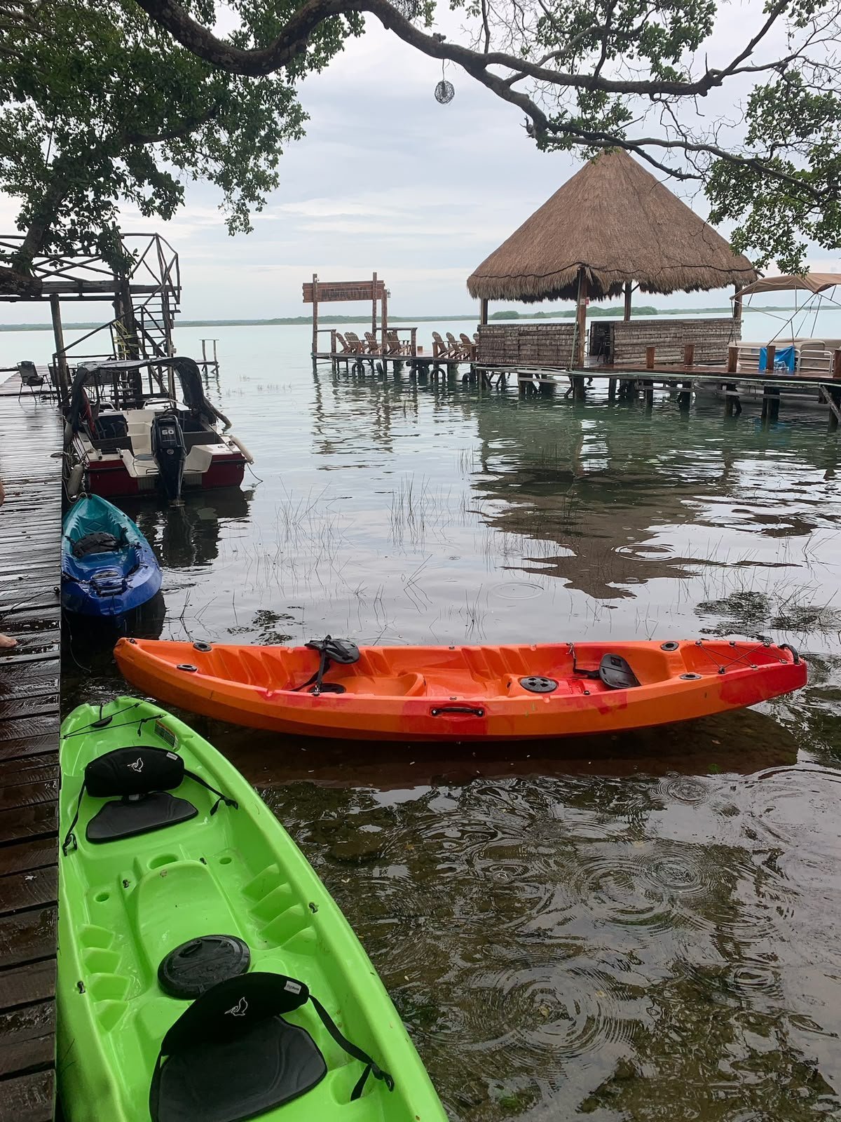 Kayaks de colores amarrados en el muelle con palapa y lancha al fondo