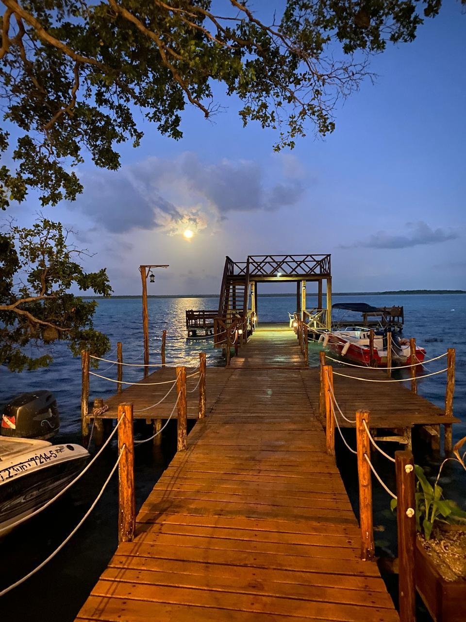 Muelle de madera del hotel con barcos y palapa iluminada al atardecer sobre la laguna