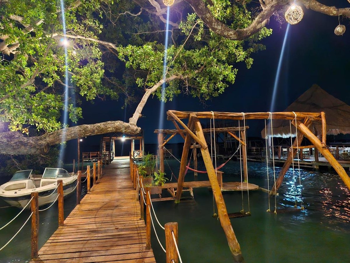 Muelle iluminado de noche con pasarela de madera hacia la palapa colgando sobre la laguna